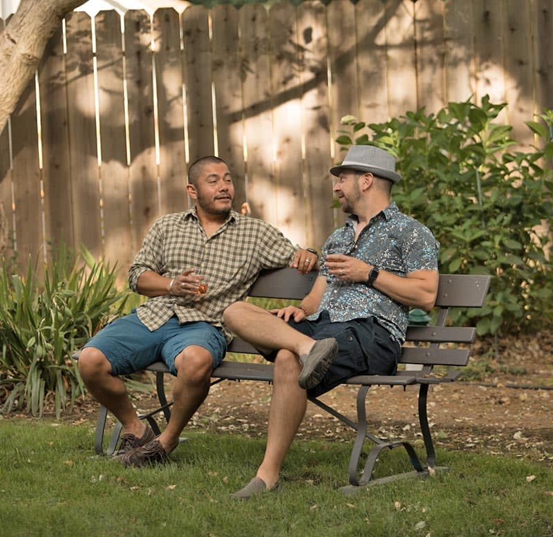 Two men share a relaxed conversation on a park bench, each holding a drink.