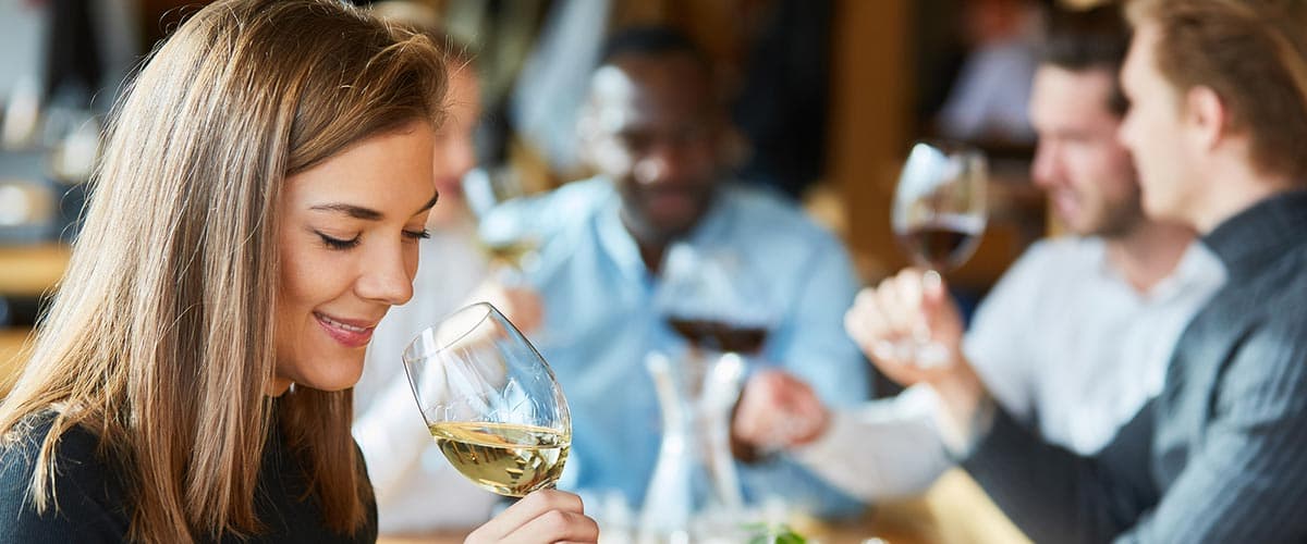 A woman smiles while holding a glass of white wine at a dinner table with friends.