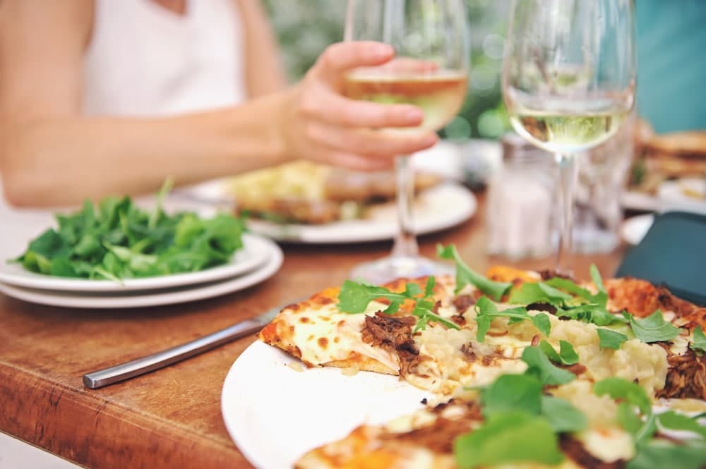 A table setting features a plate of pizza topped with greens, a side of salad, and glasses of white wine.