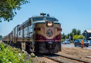 A vintage train approaches a crossing on a sunny day, surrounded by greenery and vehicles.