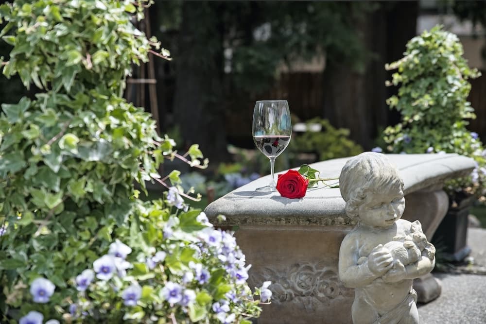A glass of red wine and a single red rose rest on a stone bench surrounded by lush greenery and blooming flowers.