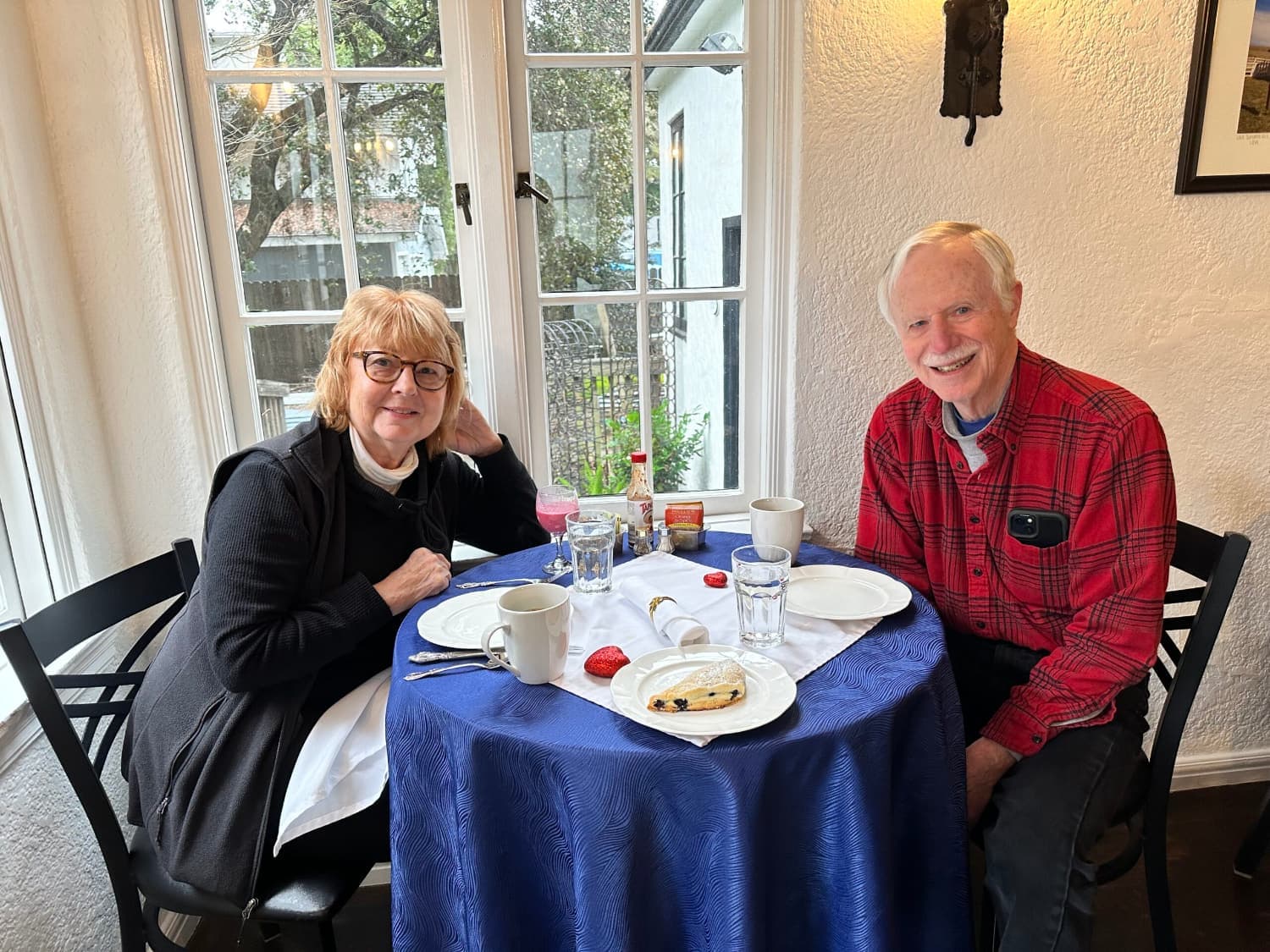 Two smiling individuals enjoy breakfast at a table set with coffee and pastries by a window.
