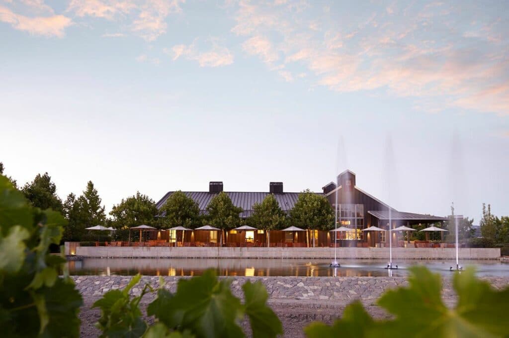 A modern building with a water feature, surrounded by greenery and under a pastel sky.