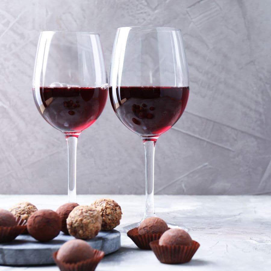 Two glasses of red wine beside assorted chocolate truffles on a grey surface.