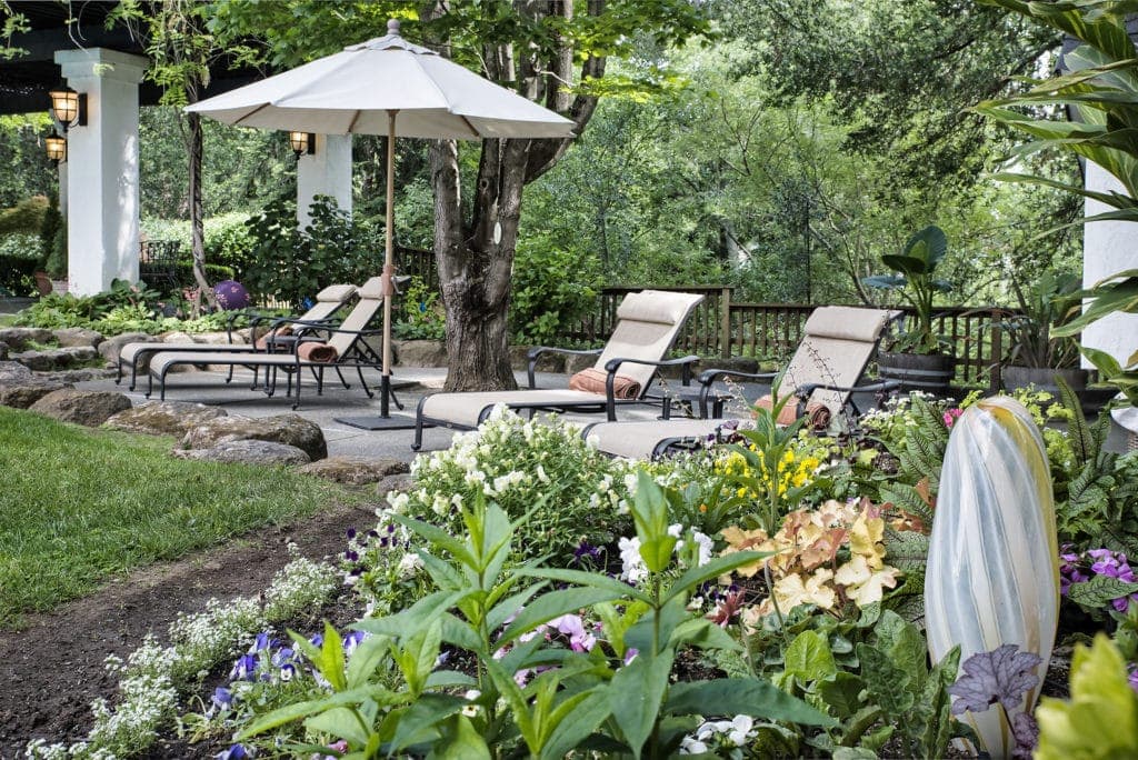 Lounge chairs under an umbrella surrounded by a lush garden.