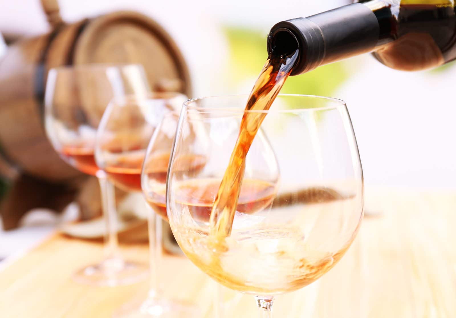 A wine bottle pours red wine into a glass, surrounded by three additional glasses on a wooden surface.