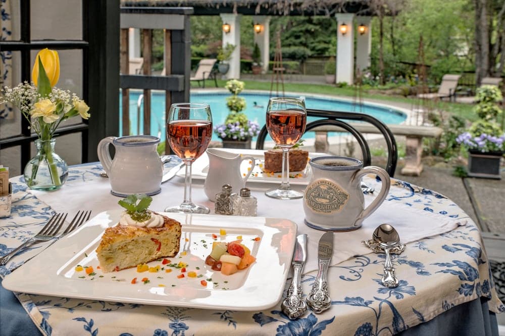 A beautifully set table features a dessert plate, two glasses of rosé wine, a coffee pot, and a view of a pool surrounded by greenery.