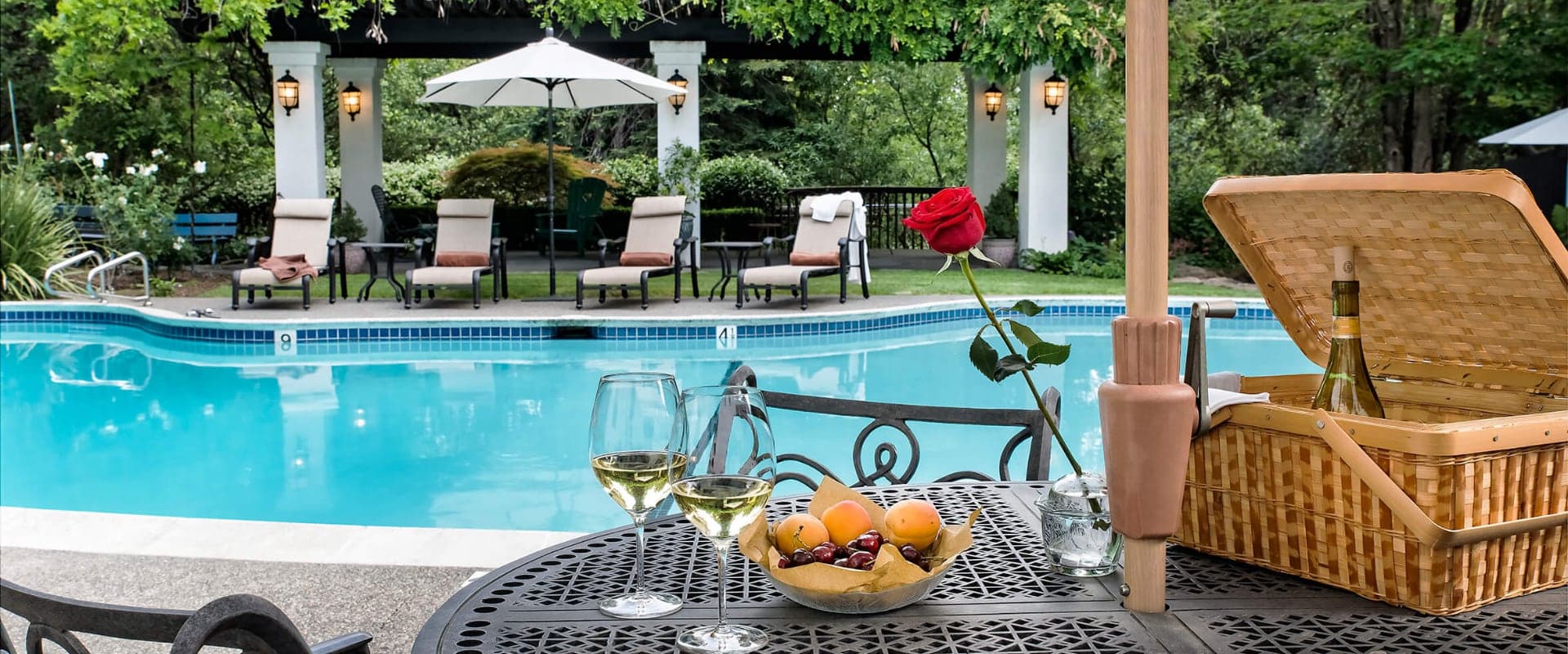 A table with fruit, wine glasses, and a picnic basket overlooks a serene swimming pool surrounded by lounge chairs and greenery.