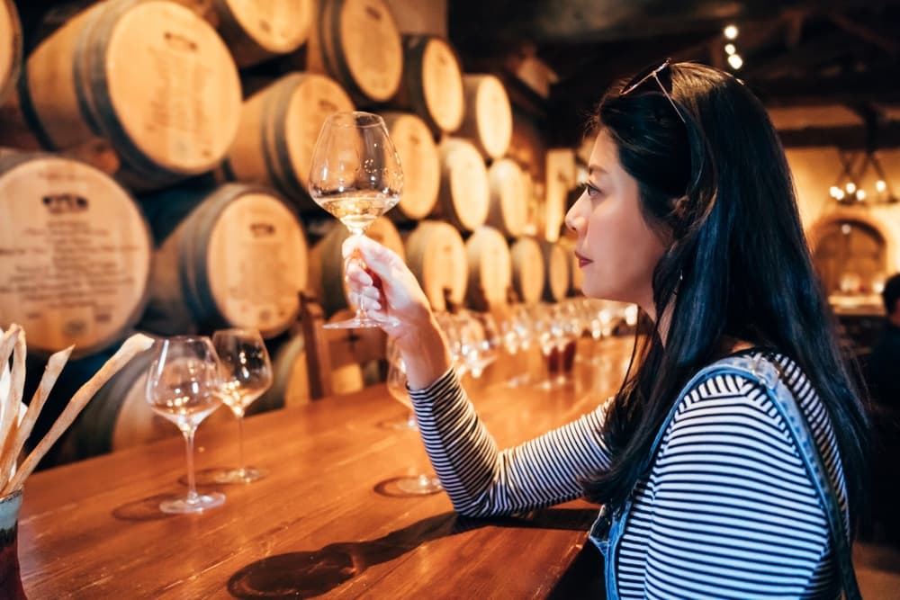 A woman inspects a glass of white wine while sitting at a wooden bar in a winery surrounded by wine barrels.