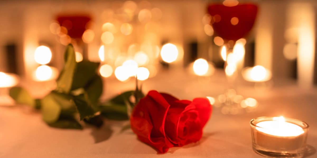 A red rose lies on a table surrounded by candles and glasses, creating a romantic atmosphere.