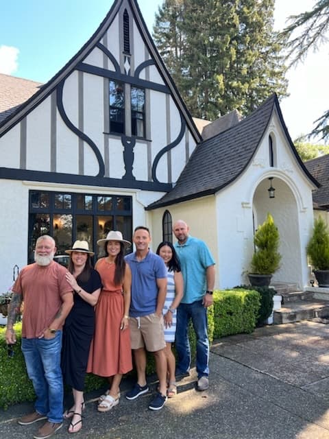 A group of six people stands in front of a charming, decorated house surrounded by greenery.