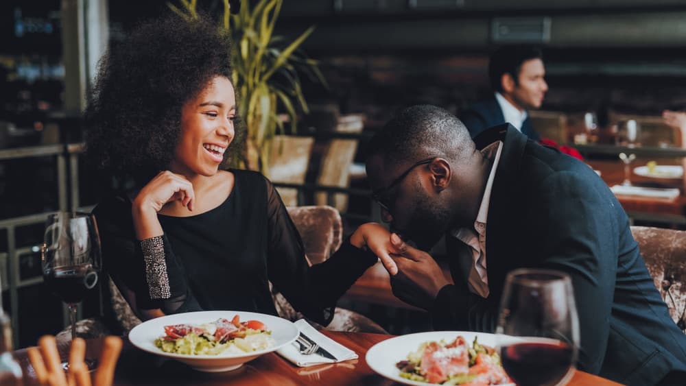 A couple shares a romantic moment at a restaurant, with one partner kissing the other's hand while they smile at each other, food and wine on the table.