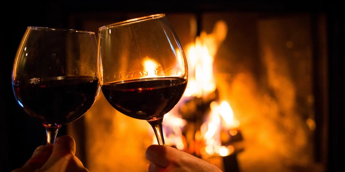 Two glasses of red wine clink together in front of a cozy fireplace.