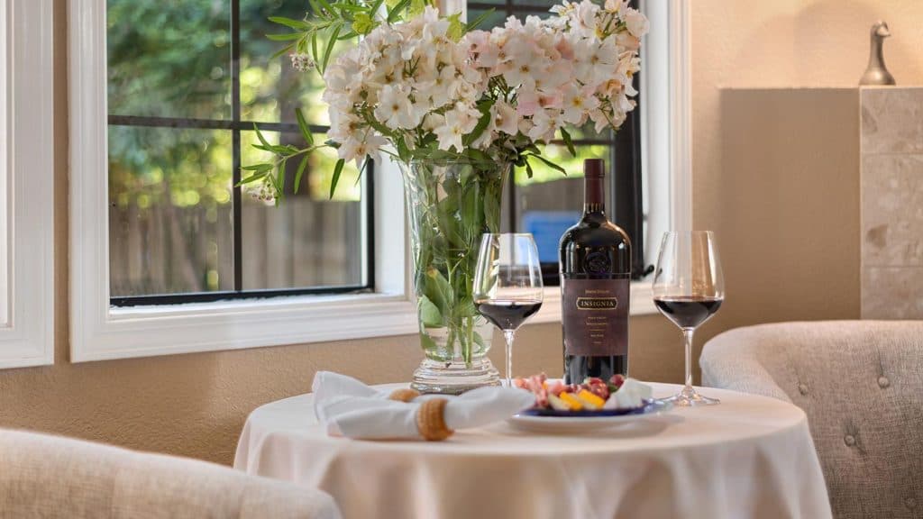 A cozy table setting with a vase of flowers, a bottle of wine, two glasses, and a plate of snacks by a window.