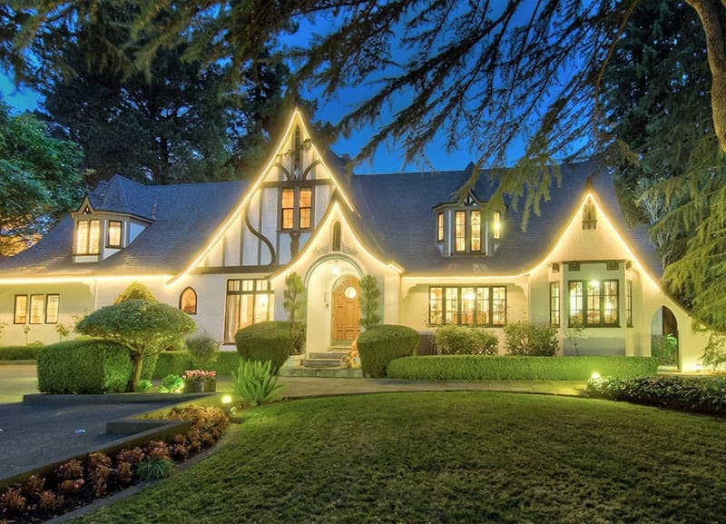 A beautifully lit, multi-gabled house surrounded by lush greenery at dusk.