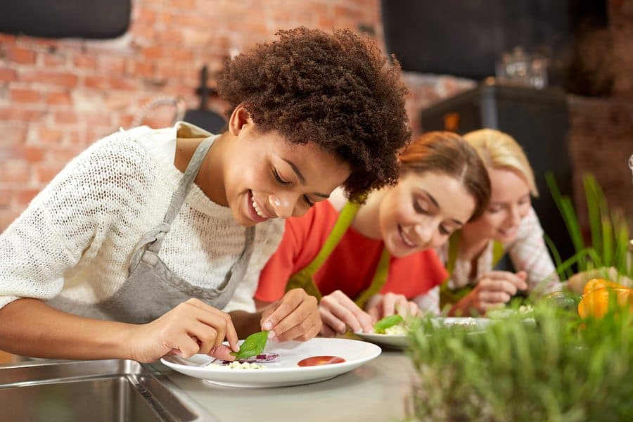 Three women joyfully arrange food on plates in a kitchen setting.