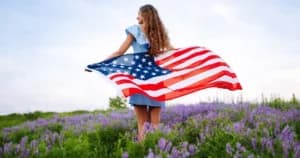 A girl in a blue dress stands in a field of purple flowers, holding a waving American flag. A girl in a blue dress stands in a field of purple flowers, holding a waving American flag.
