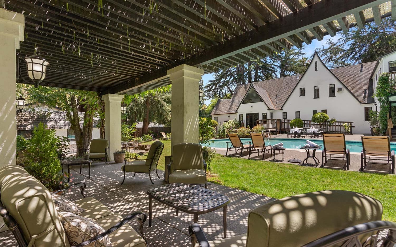 A shaded patio area with comfortable seating overlooks a pool surrounded by greenery and a white building.