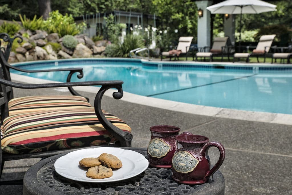 A plate of cookies and two mugs sit on a table beside a poolside chair.