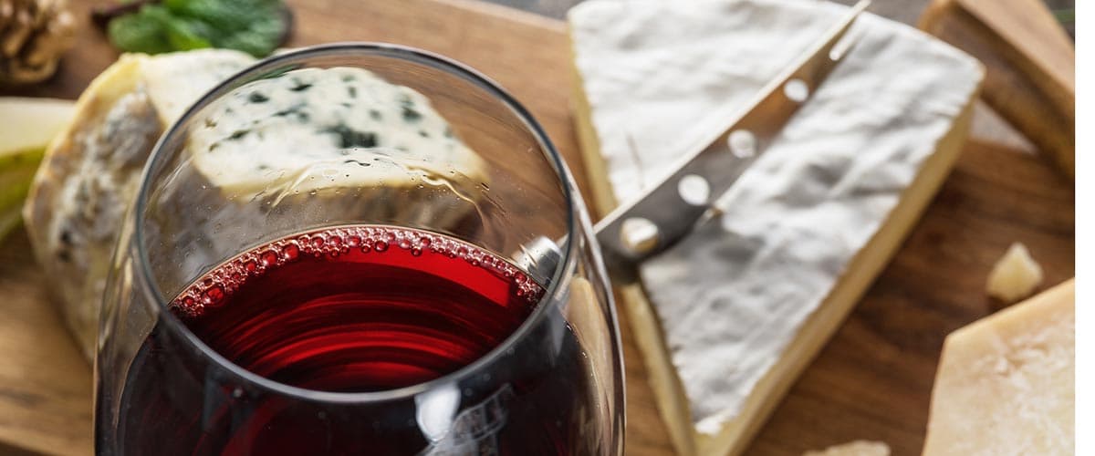 A glass of red wine sits beside an assortment of cheeses on a wooden board.