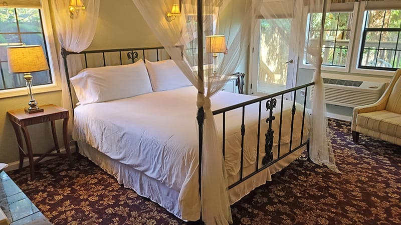 A cozy bedroom featuring a four-poster bed with white linens, two lamps, and large windows.