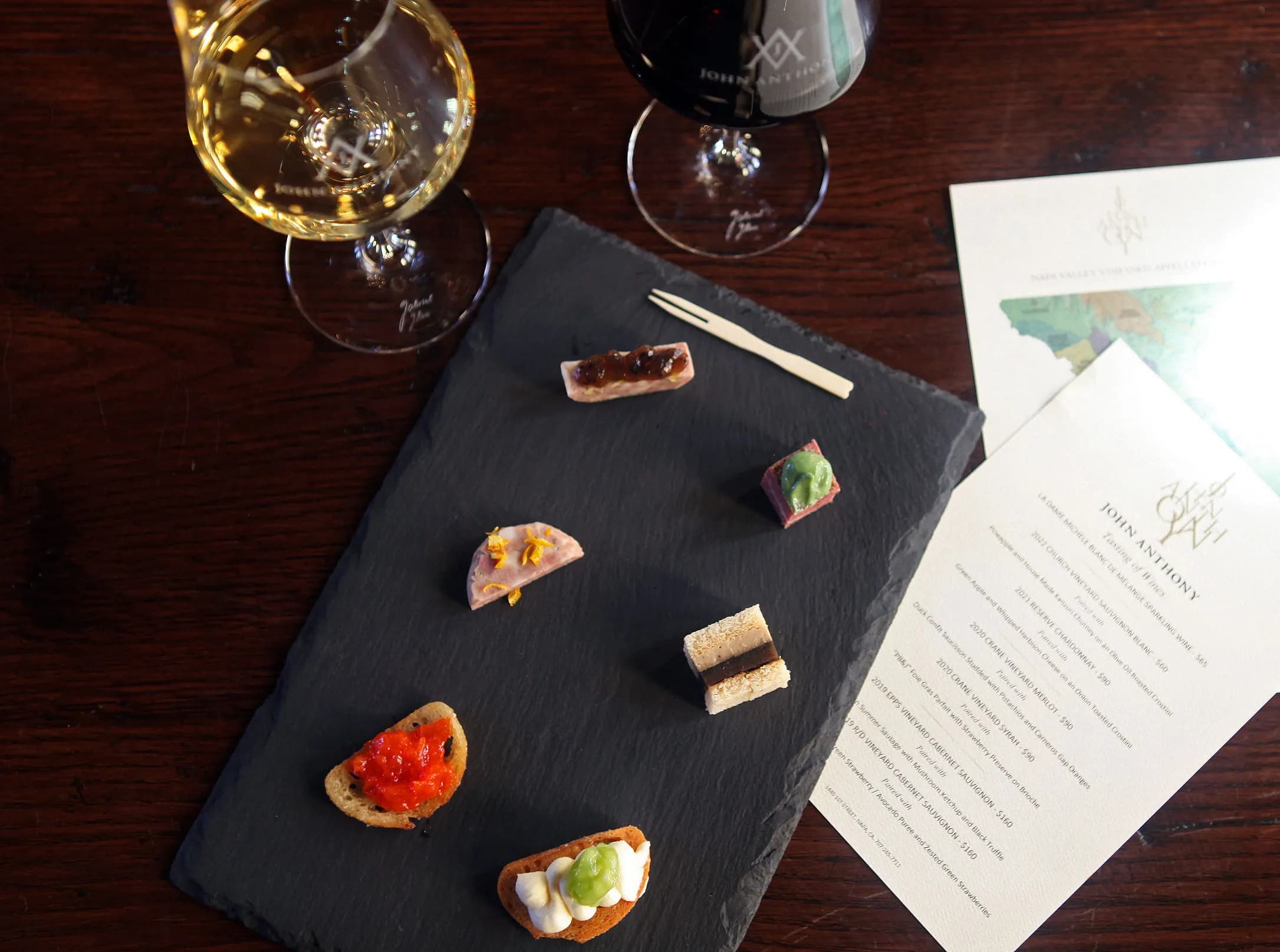 A slate platter with assorted small gourmet bites, accompanied by two glasses of wine and a menu.