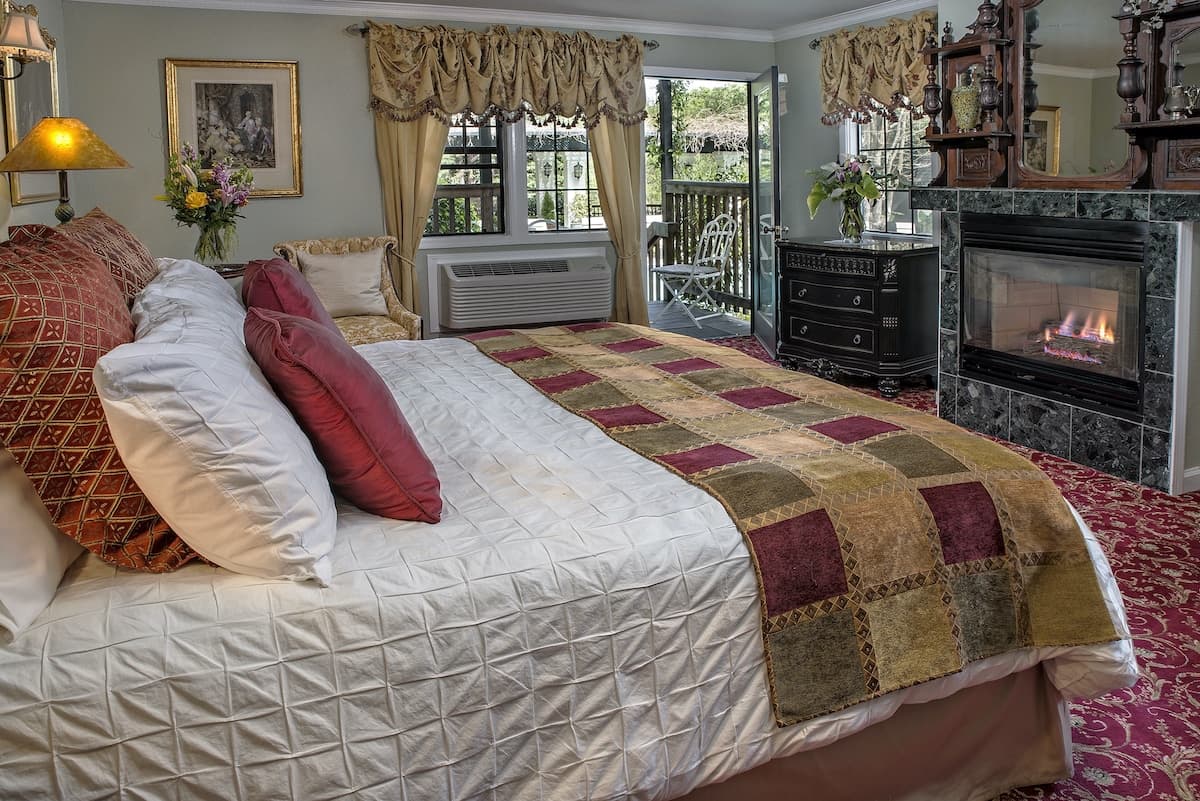 A cozy bedroom with a patterned quilt, plush pillows, and a fireplace, featuring large windows that open to a patio.