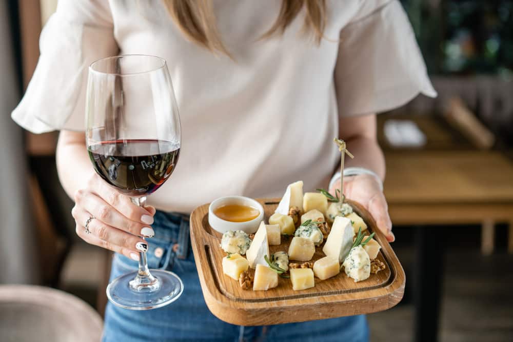 A person holds a glass of red wine in one hand and a wooden platter of assorted cheeses in the other.