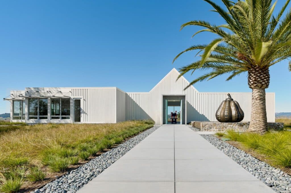 A modern, white-clad building with a pathway leading to its entrance, surrounded by green landscaping and palm trees.