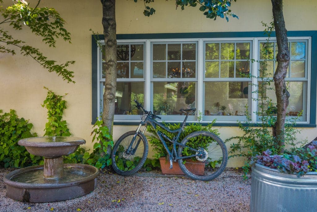A black bicycle leans against a wall near a fountain and a window with greenery surrounding it.