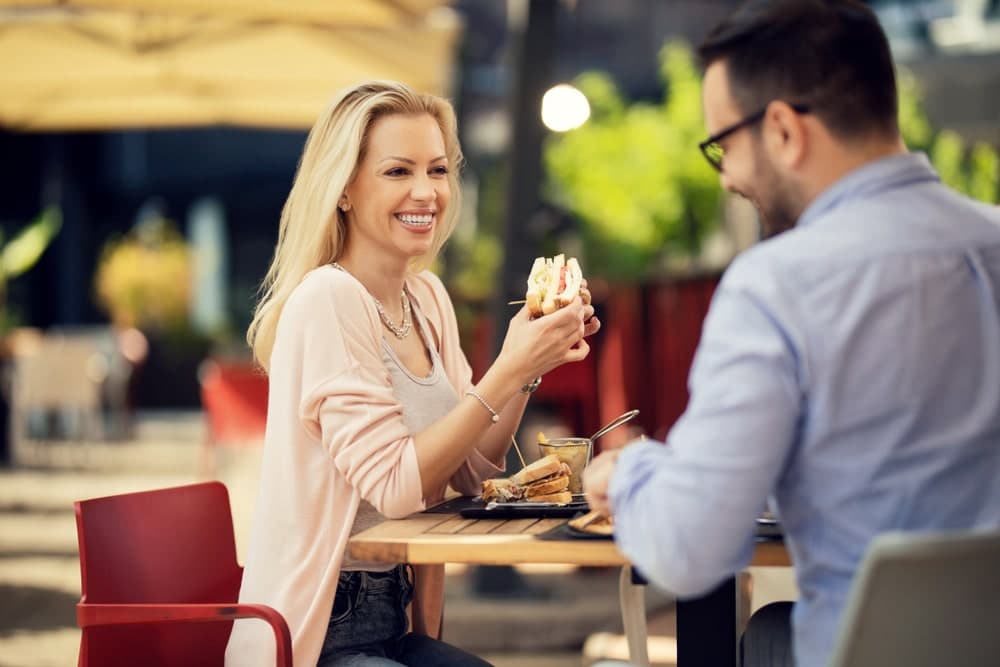 A smiling woman enjoys a sandwich while sitting at an outdoor table with a man.