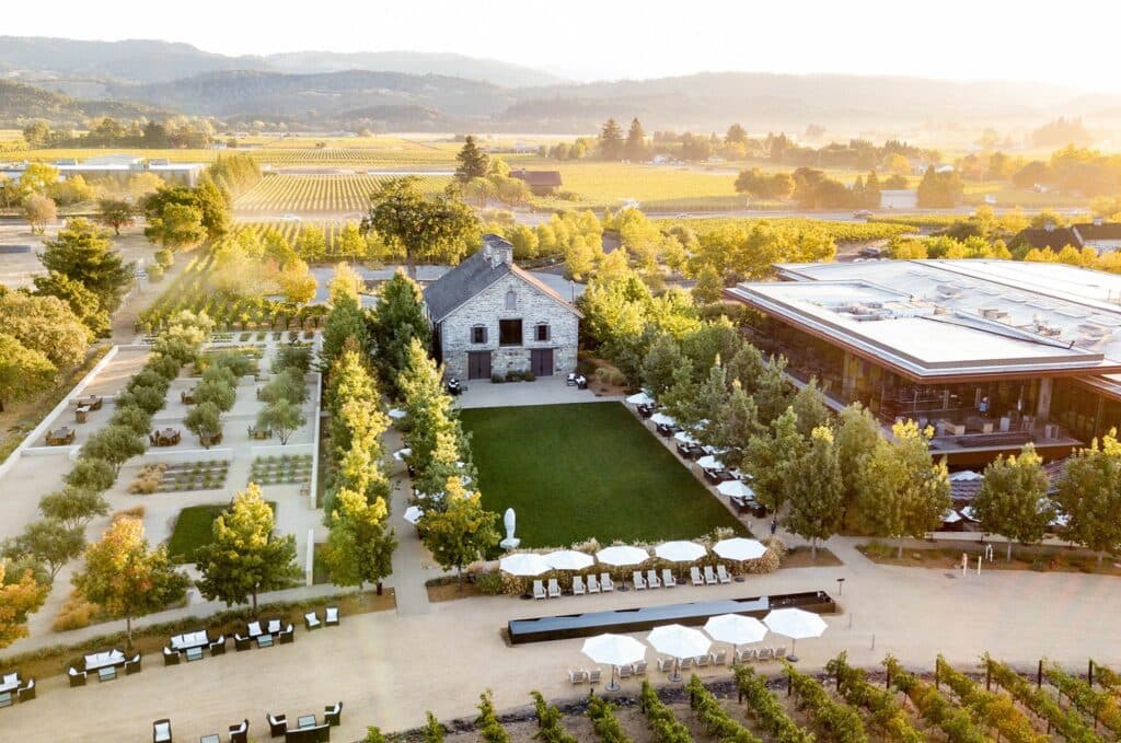 Aerial view of a vineyard resort with a modern building, manicured gardens, and lounge areas during sunset.
