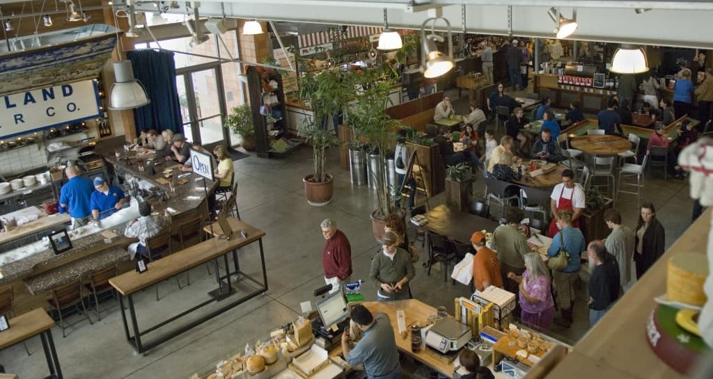 A bustling indoor market filled with customers dining and shopping, featuring communal tables and a coffee bar.
