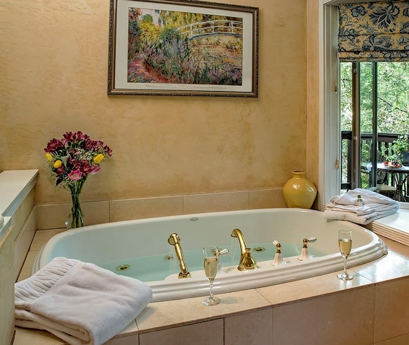 A luxurious bathroom featuring a jetted tub, fresh flowers, wine glasses, and natural light.