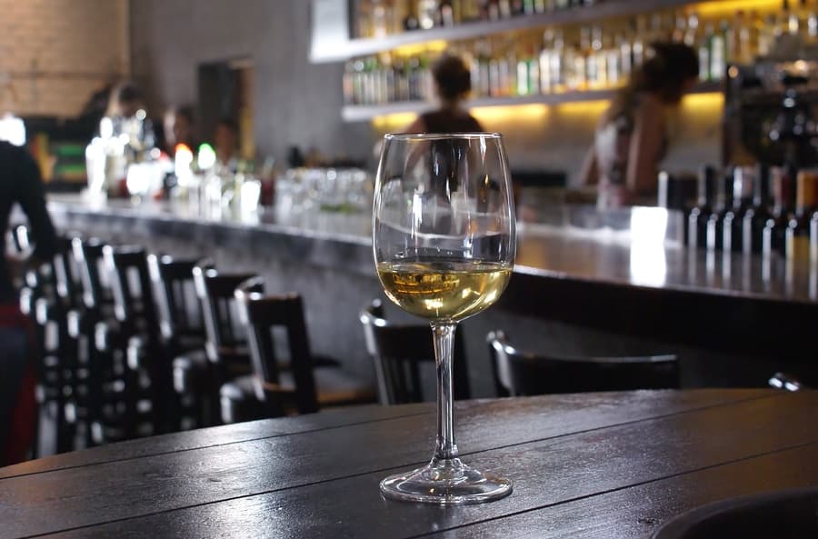 A glass of white wine sits on a wooden table in a dimly lit bar.