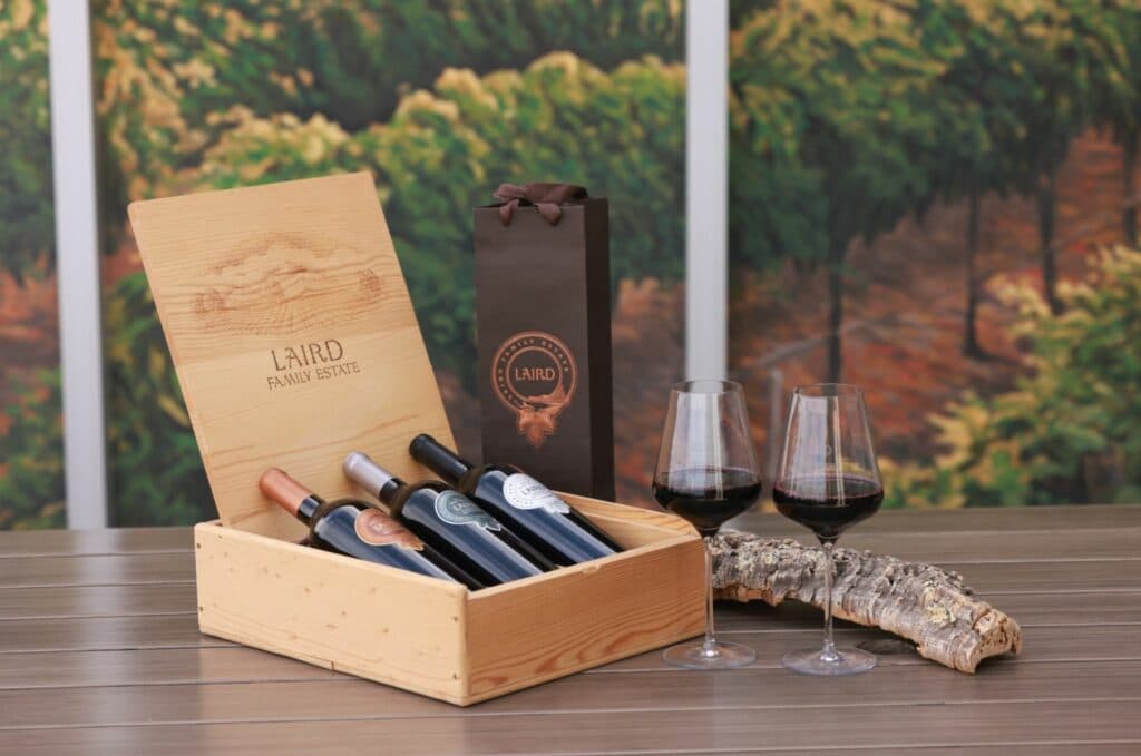 A wooden box with three wine bottles, a gift bag, and two glasses of red wine displayed on a table.