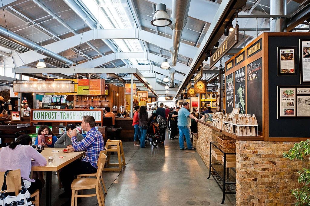 A bustling indoor food market with various stalls and patrons dining at wooden tables.