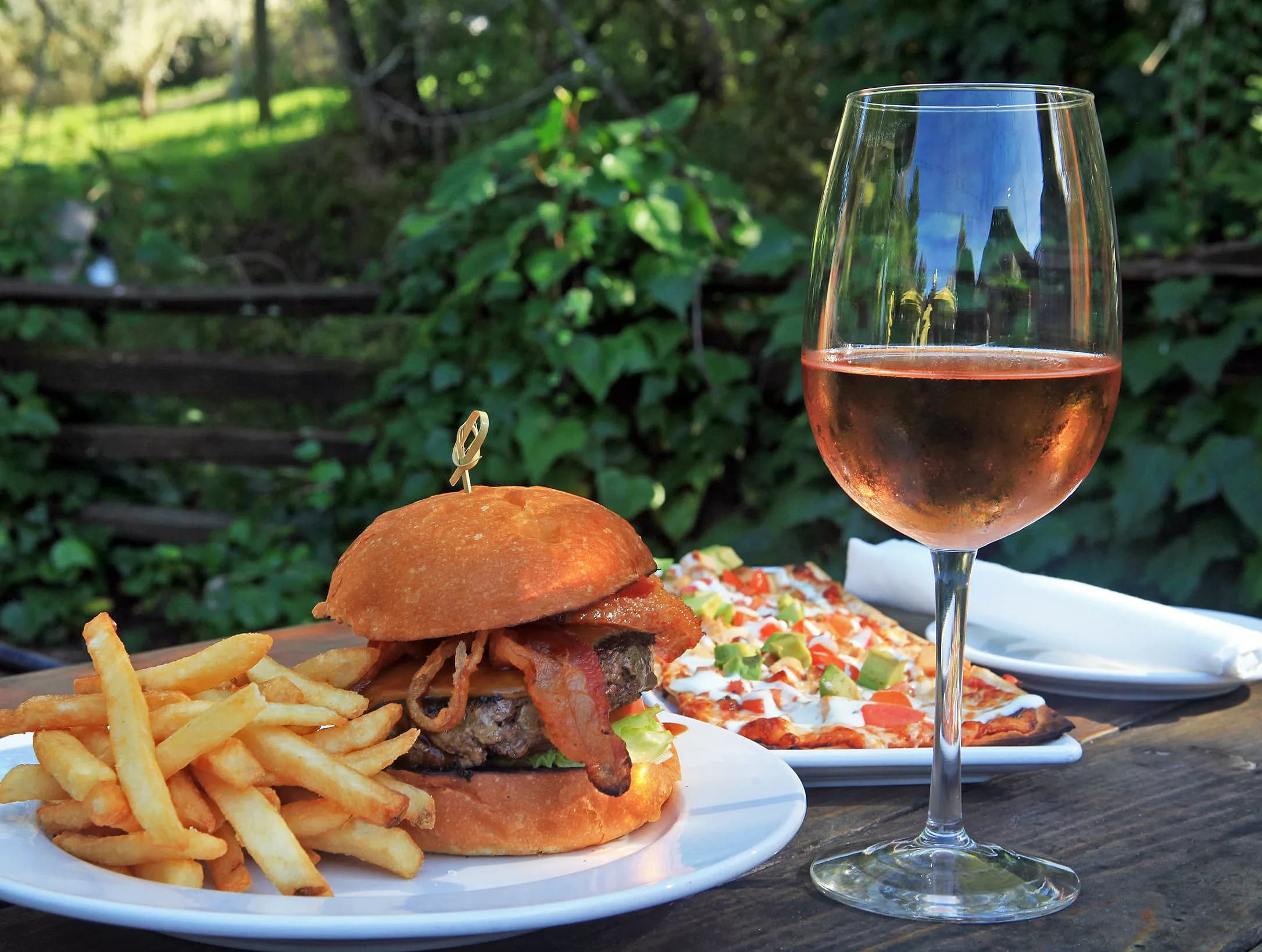 A plate of a burger with fries, a glass of rosé wine, and a pizza slice in a lush outdoor setting.