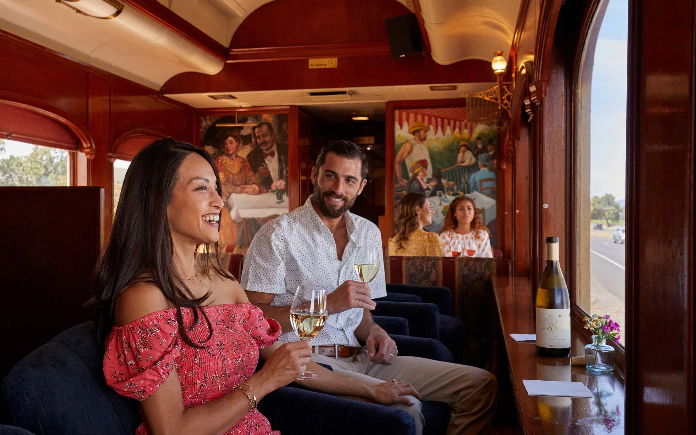A woman and a man enjoy drinks on a luxurious train, with vibrant artwork and a scenic view outside the window. A woman and a man enjoy drinks on a luxurious train, with vibrant artwork and a scenic view outside the window.