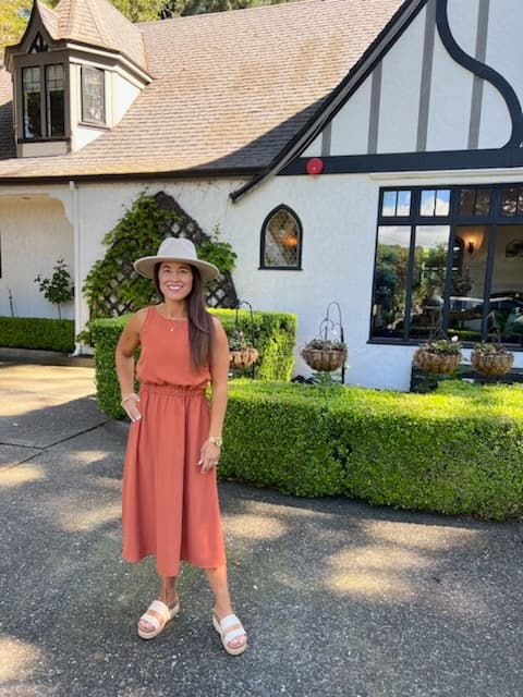 A woman in a rust-colored dress and wide-brimmed hat stands in front of a charming building surrounded by greenery.