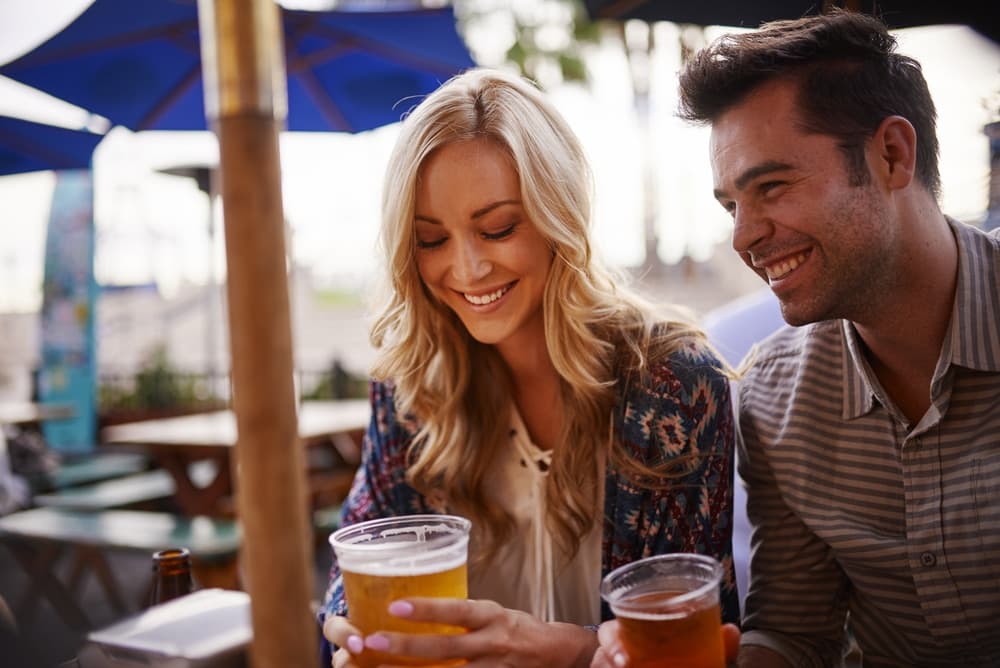 A woman and a man smile while enjoying drinks at an outdoor table under a blue umbrella.