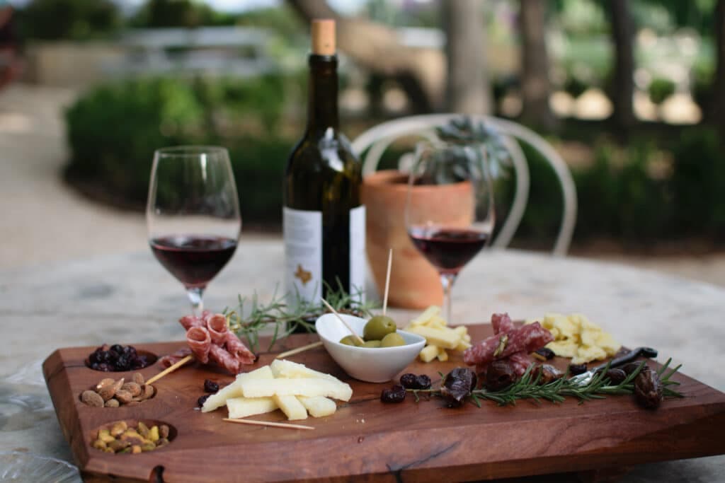 A wooden platter with various cheeses, cured meats, olives, nuts, and dried fruits, accompanied by two glasses of red wine and a bottle, set in a garden.