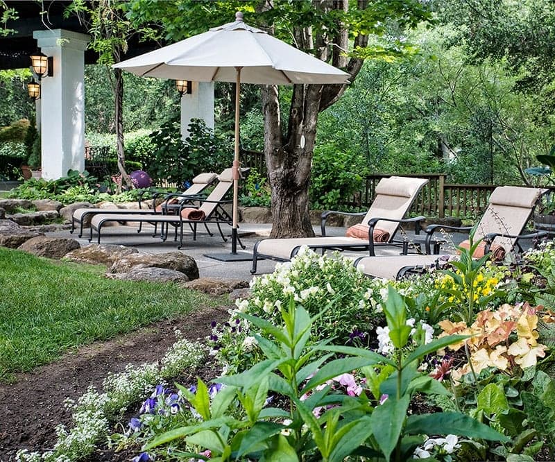Lounge chairs and an umbrella are nestled in a lush garden with colorful flowers.