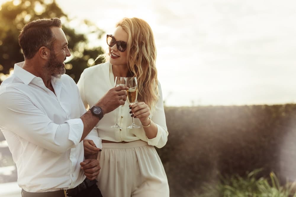 A man and woman toast with champagne outdoors, both smiling and dressed elegantly.