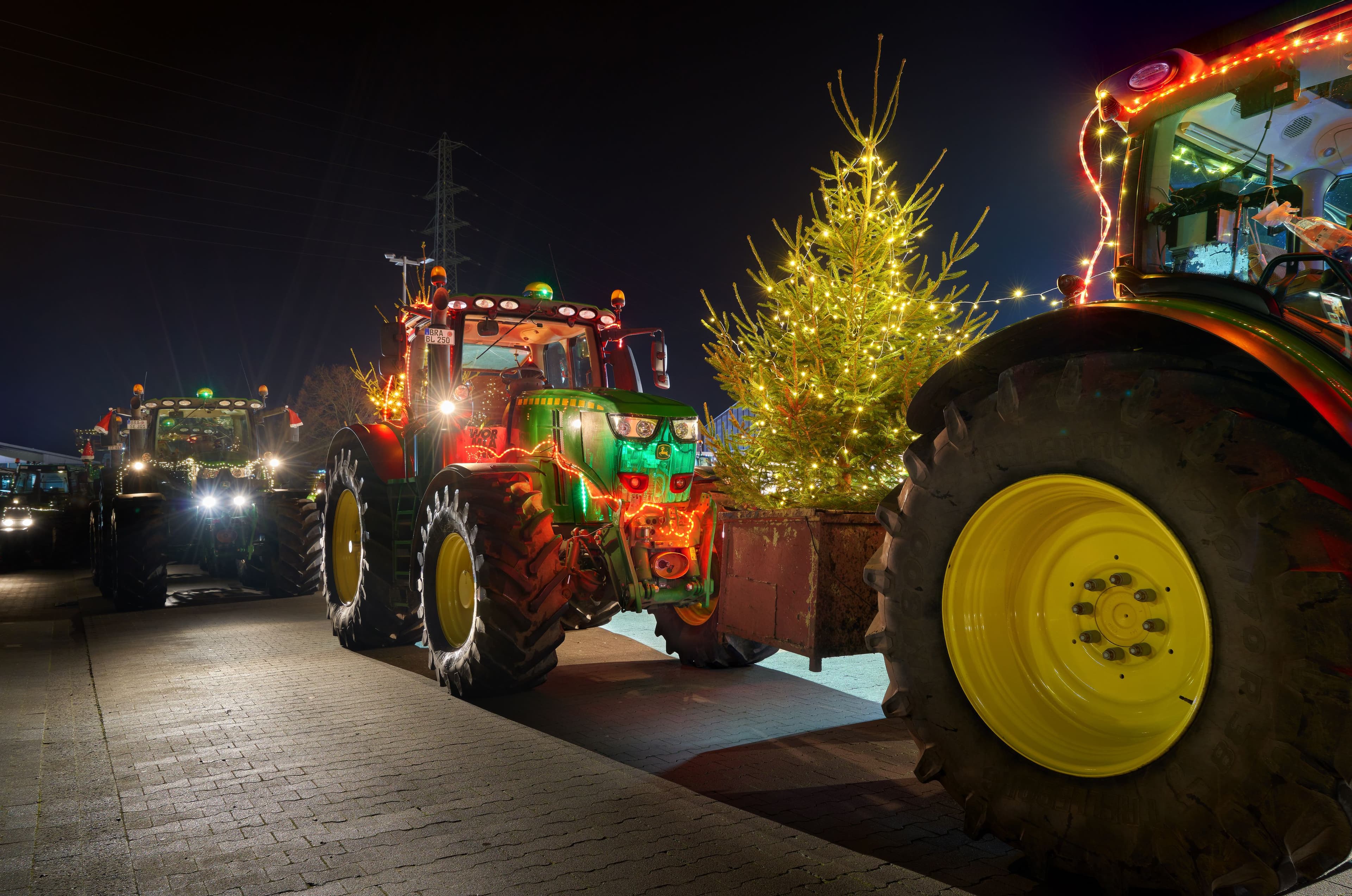 Two brightly lit tractors pulling decorative Christmas trees at night.