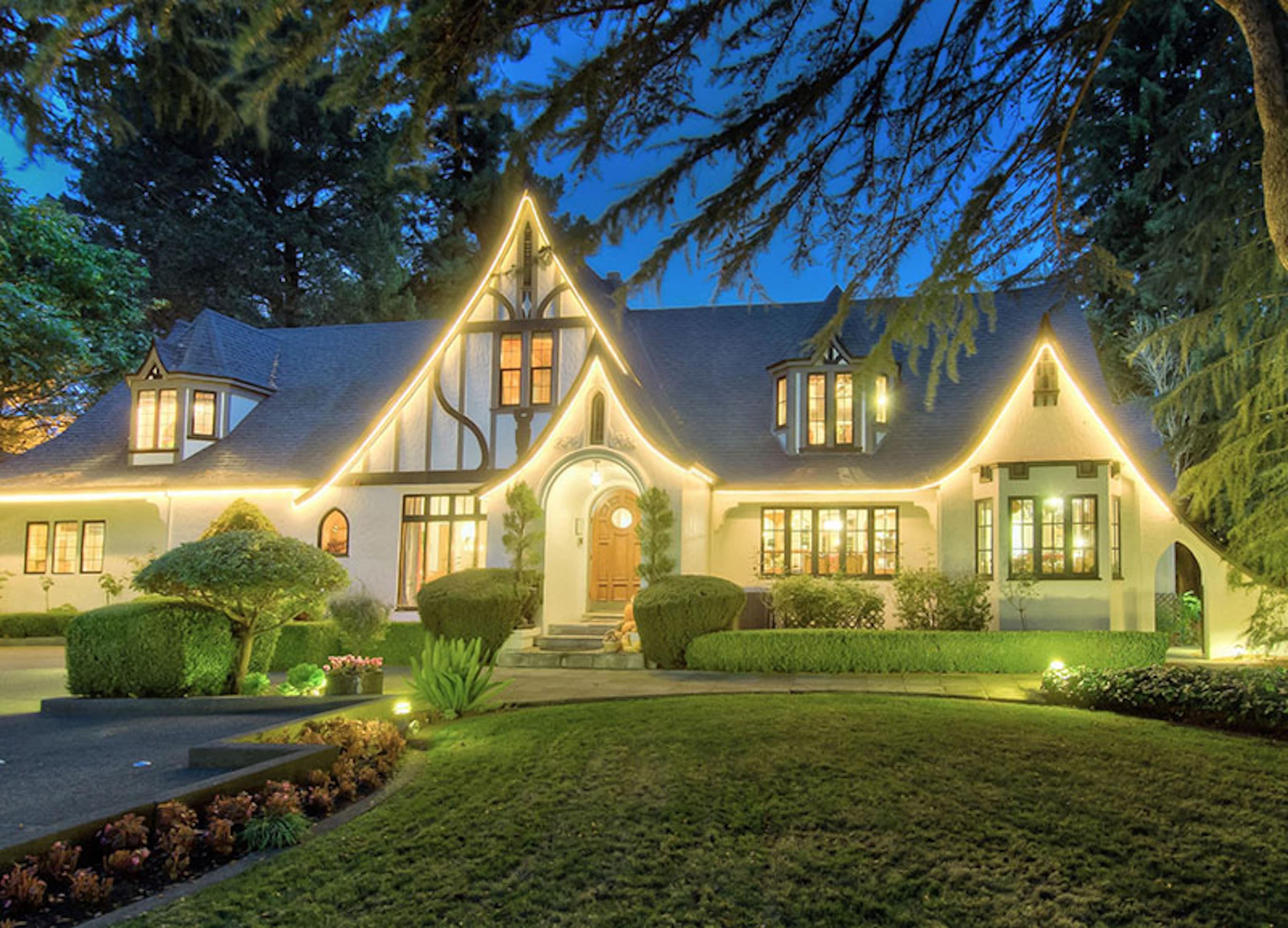 A beautifully lit, modern Tudor-style house surrounded by manicured gardens at dusk.
