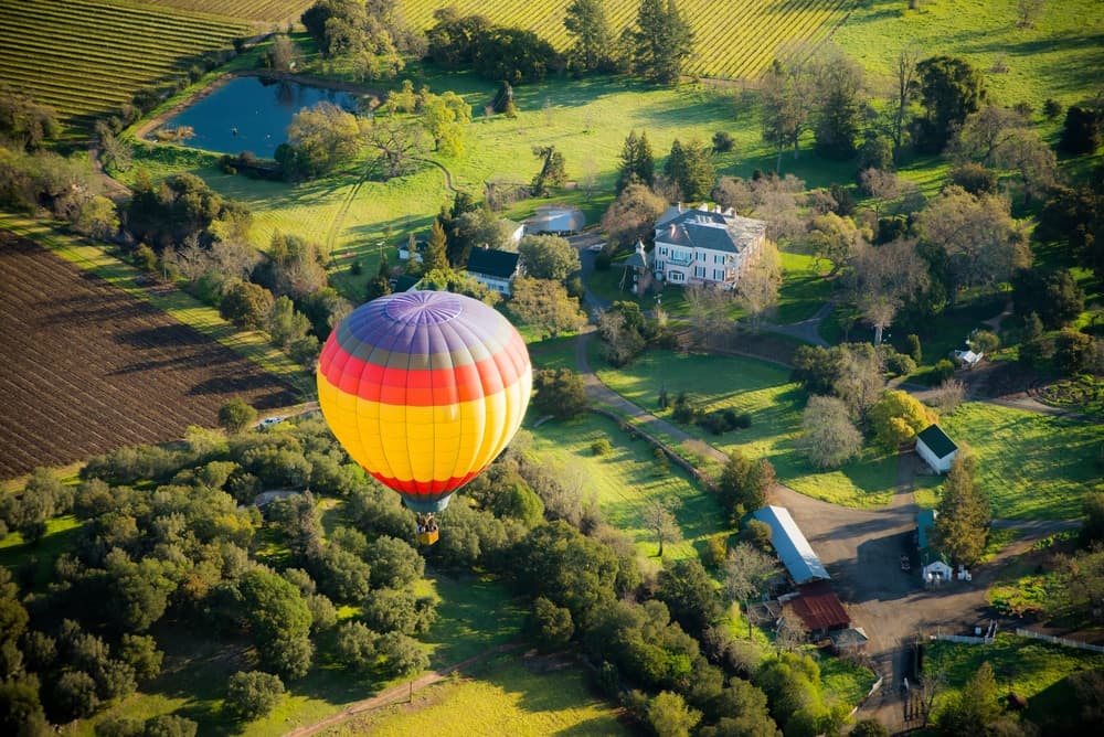 A colorful hot air balloon floats over a lush green landscape with a house and pond below.