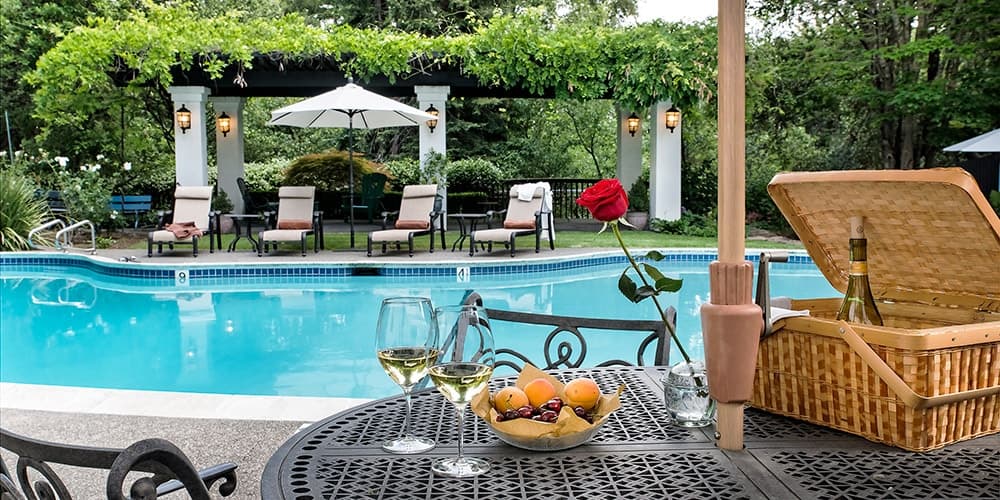 A sunny outdoor pool area with loungers, an umbrella, and a table set with wine glasses, a picnic basket, and a bowl of fruit.