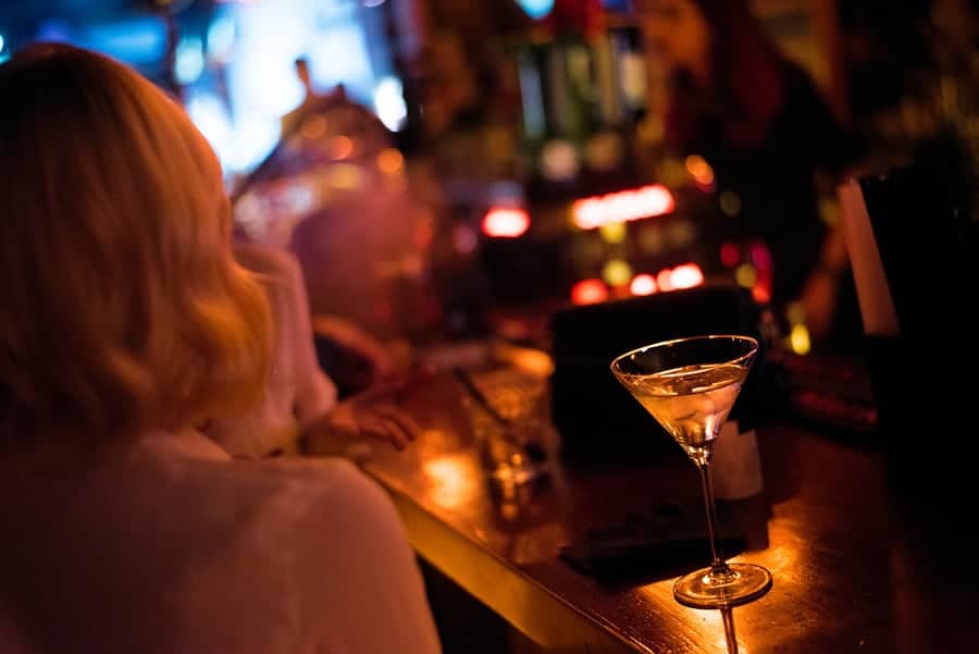 A cocktail glass sits on a dimly lit bar while a person with short hair is seated nearby.