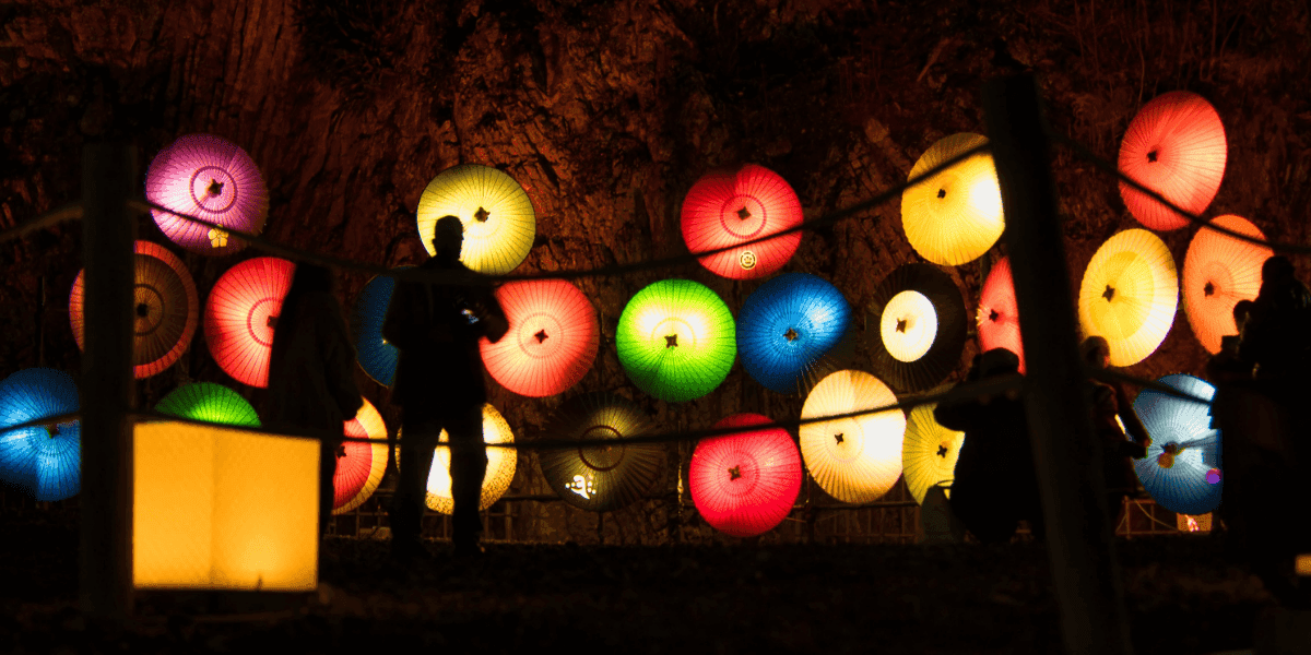 Silhouettes of people stand in front of colorful illuminated lanterns on a dark background.